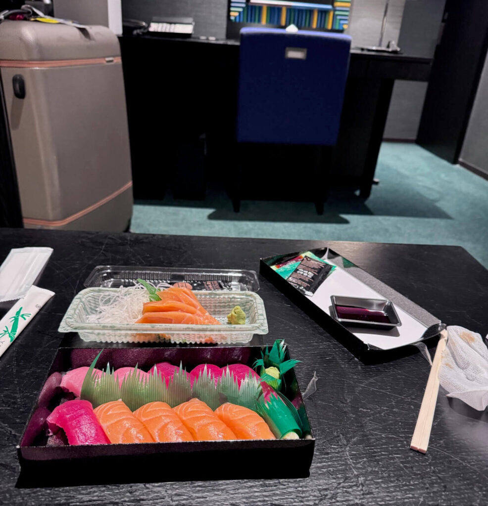 Takeout tuna and salmon nigiri and salmon sashimi on a hotel room table with a suitcase in the background