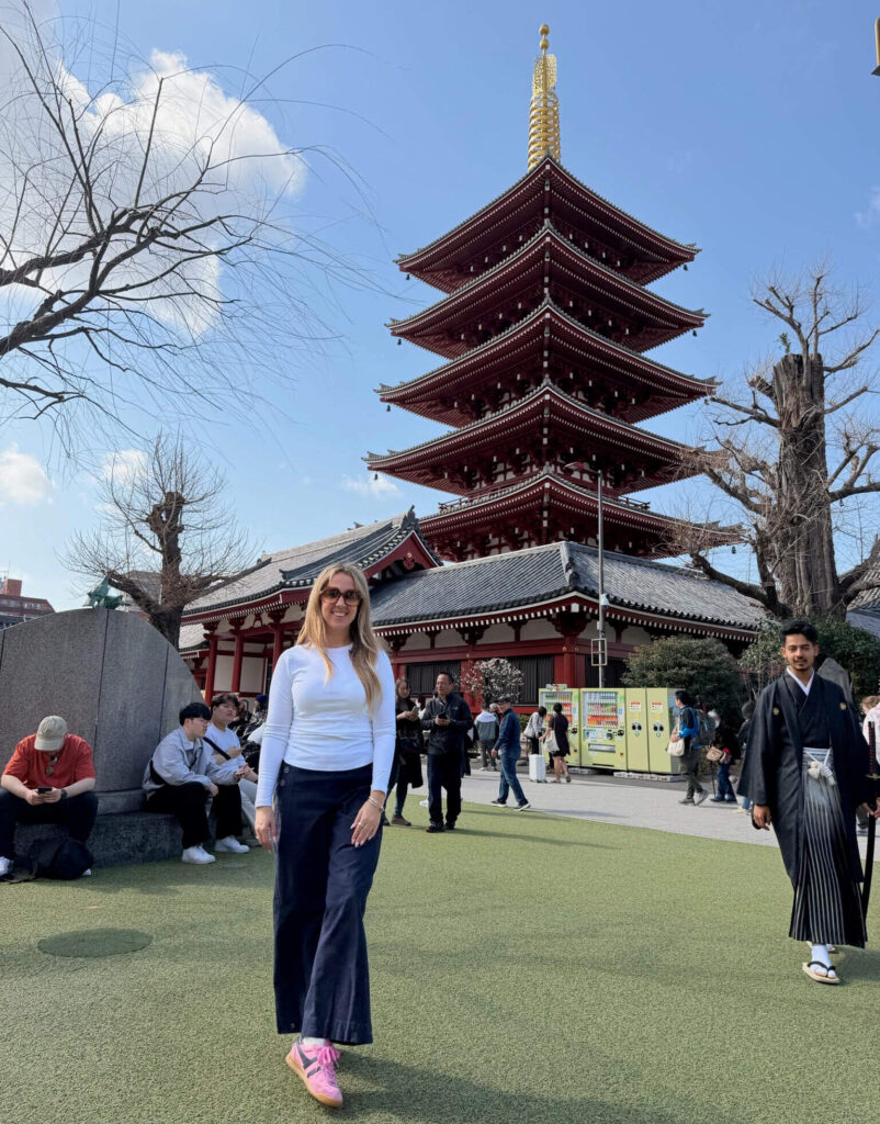 Woman posing in front of the five-story red pagoda at Senso-ji Temple in Tokyo, with a man in traditional dress walking past