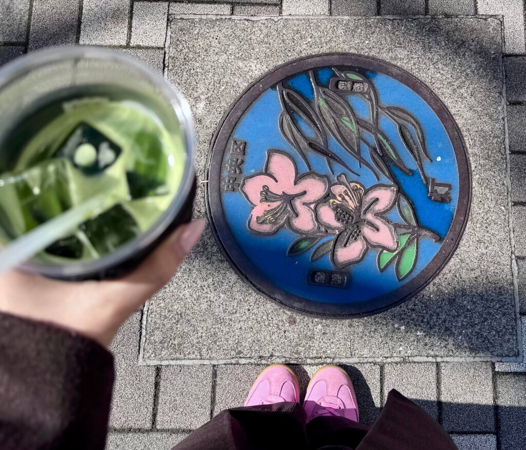 Blue and pink Chuo Ward manhole cover with azalea blossoms beside a hand holding an iced matcha drink