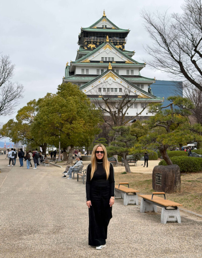 Woman standing on a gravel path in front of Osaka Castle with its green tiled roofs and gold tiger decorations.