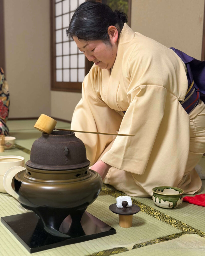 Woman in a cream kimono kneeling on tatami beside an iron kettle and bamboo ladle during a tea ceremony in Tokyo