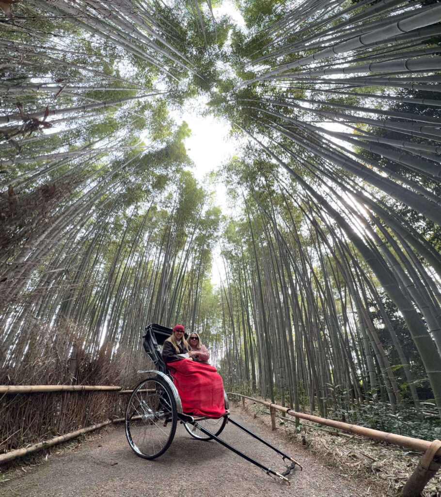 Two women riding in a rickshaw under a tall canopy of bamboo in the Arashiyama Bamboo Forest